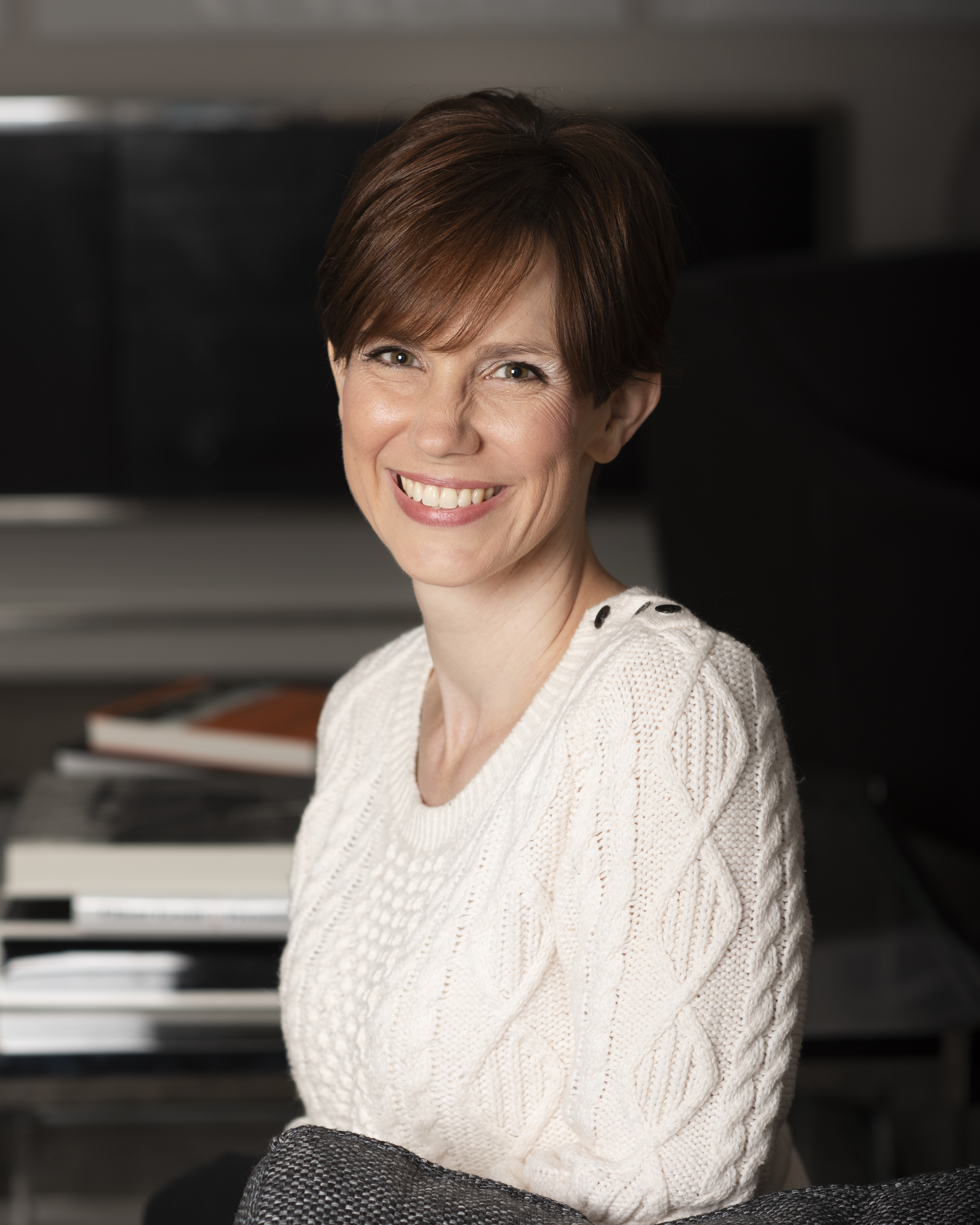 A white woman with short brown hair wearing a cable knit sweater sits facing the camera, in front of a stack of books