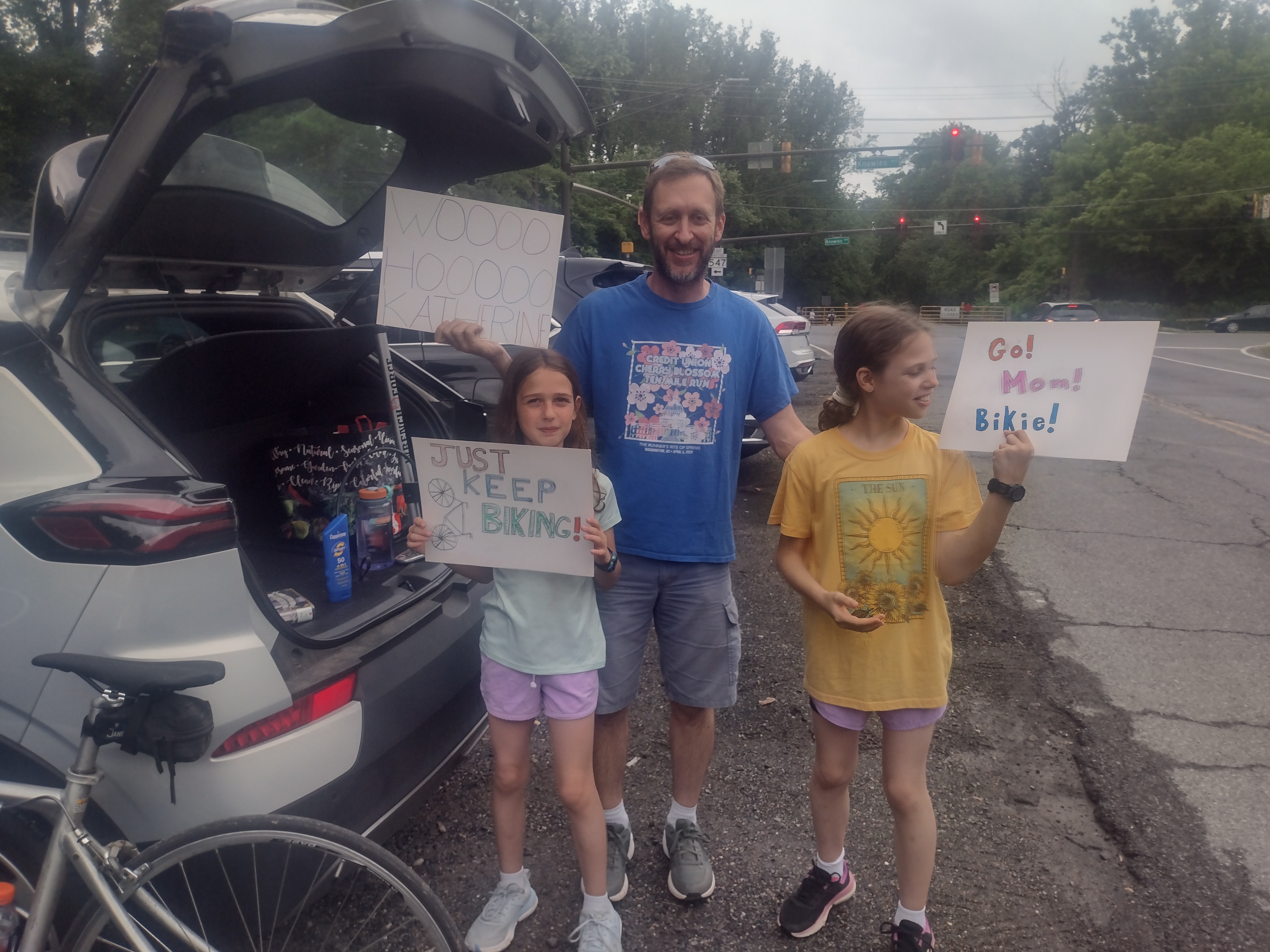 A man and two daughters standing at the back of a car with the hatch open. All three are holding handmade signs