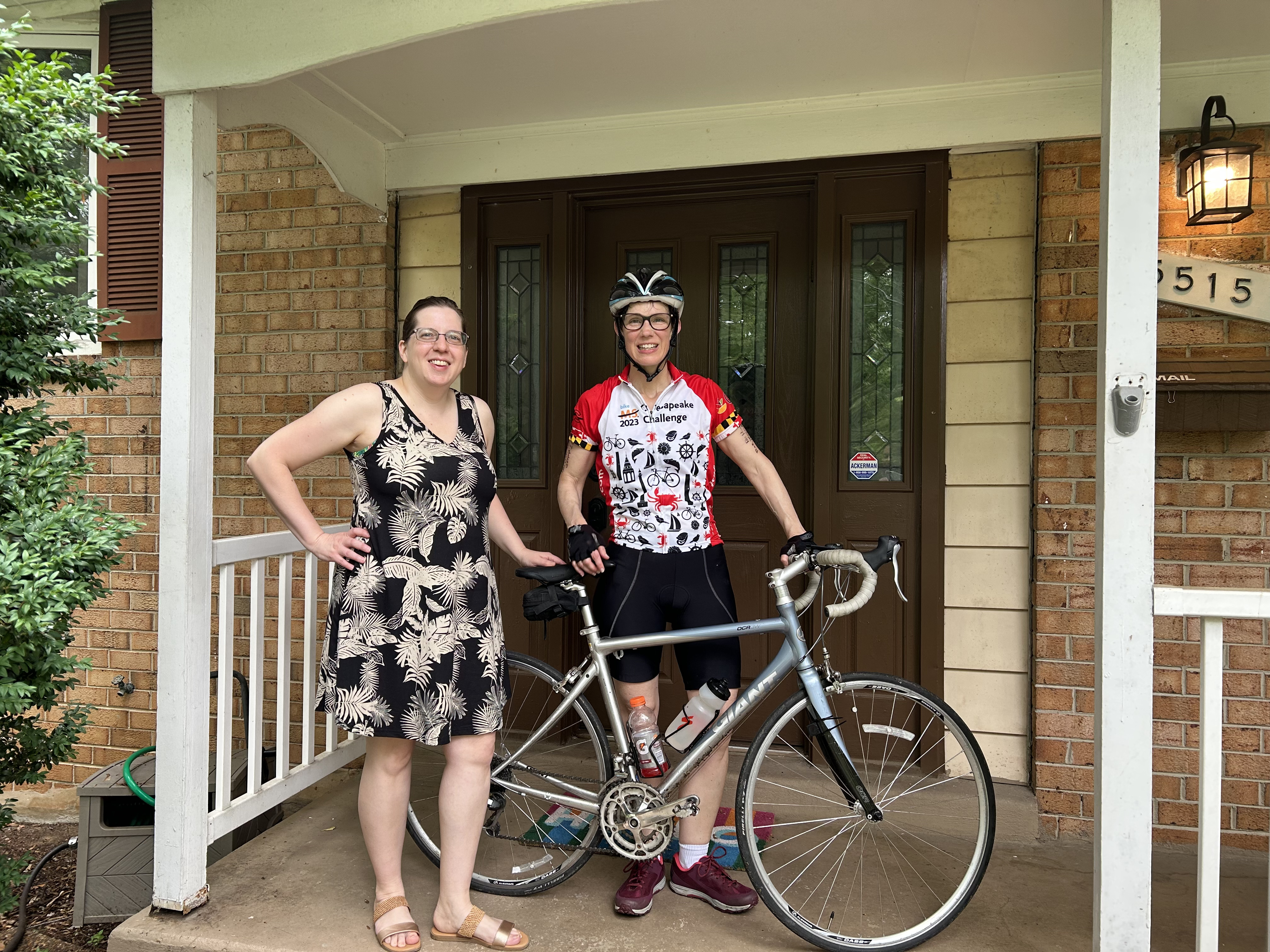 Photo of two women. One is wearing a brown and white sundress. The other is in full biking gear. A road bike is between them.