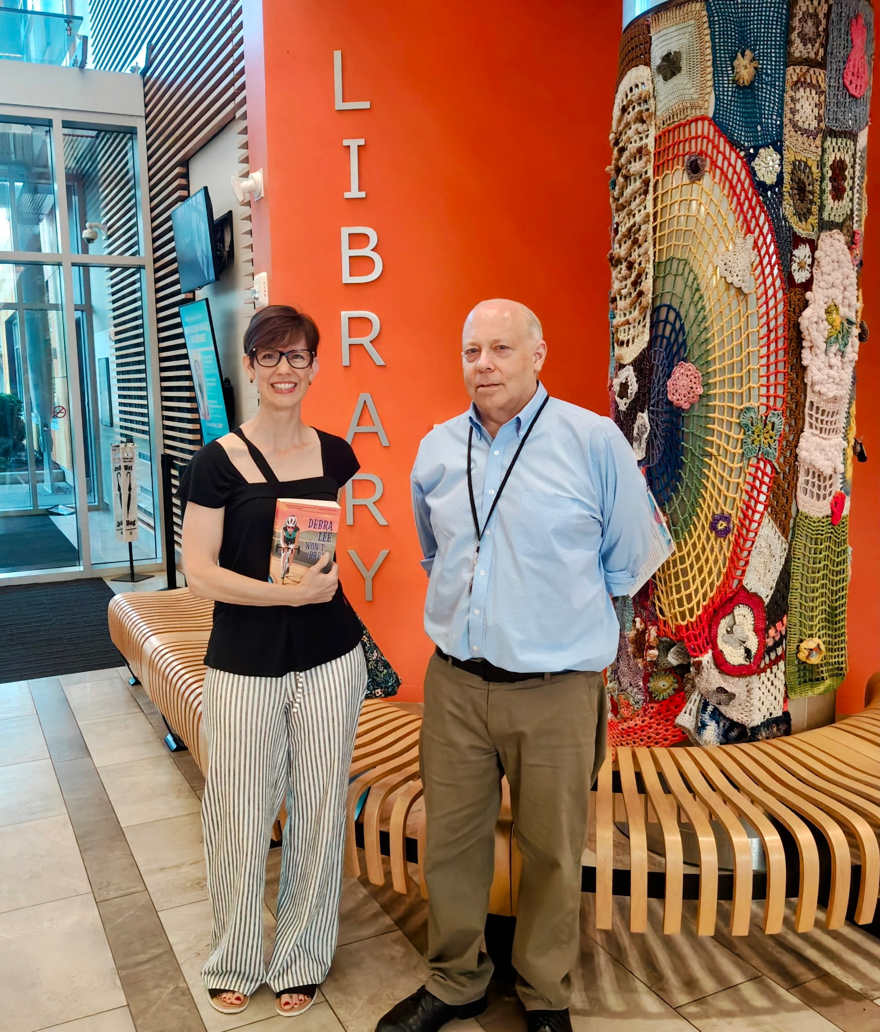 Photo of a white woman in a black shirt posing next to a white man in a light blue shirt in front of an orange wall. The word Library is visible on the wall behind them