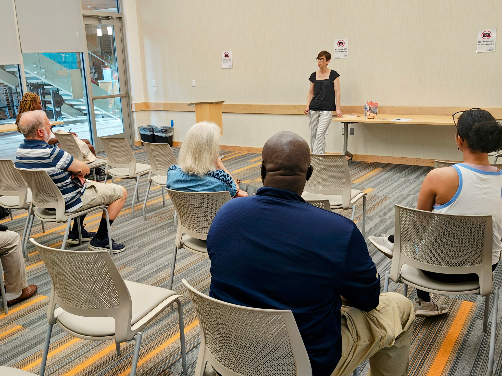 A view of a room with several people sitting in chairs facing away from the camera. They are watching a woman in a black shirt and white pants who is talking at the front of the room