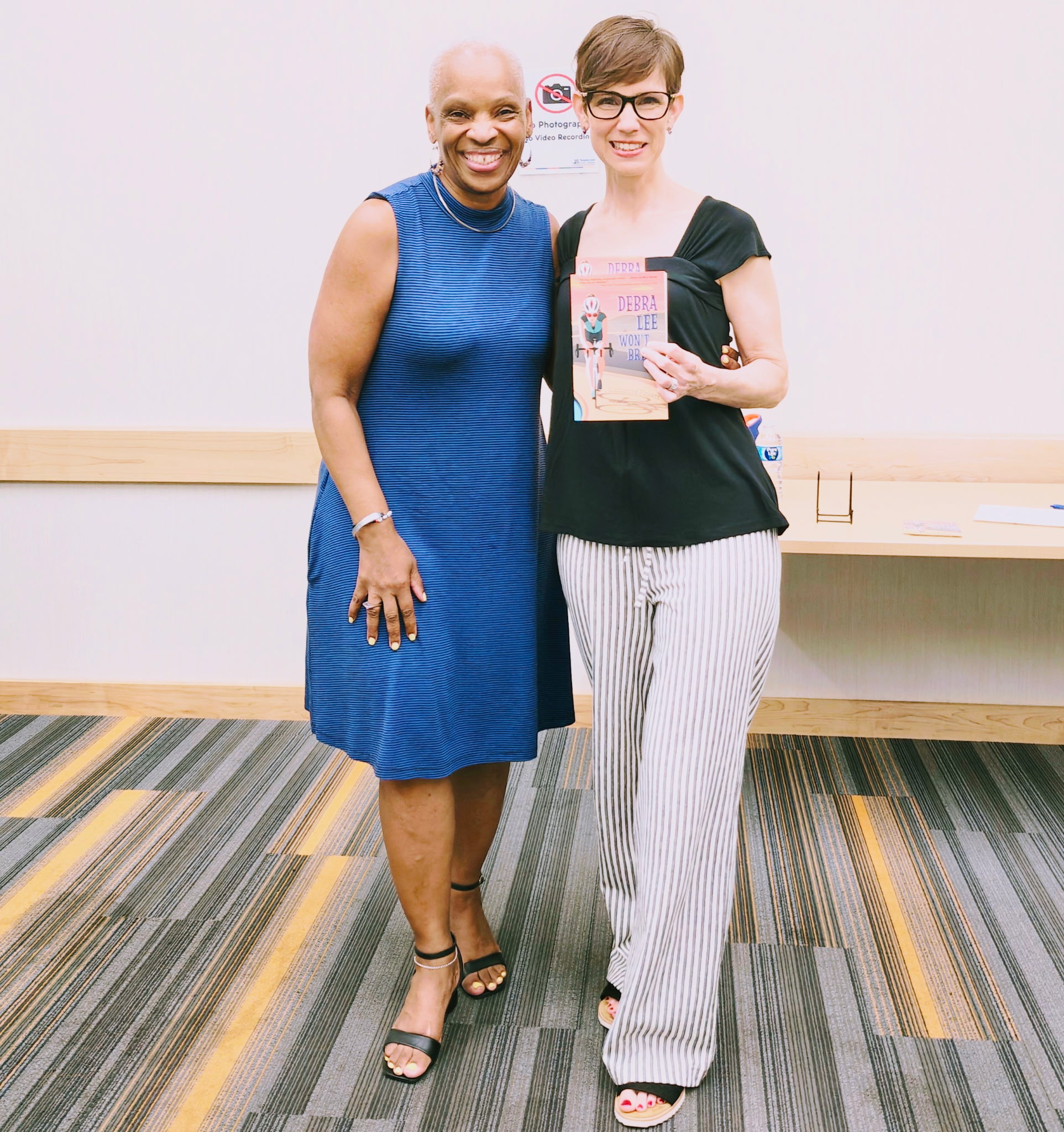 Two women pose for a picture. The woman on the right is wearing a vibrant blue dress. The woman on the left wears a black shirt with black-and-white pants