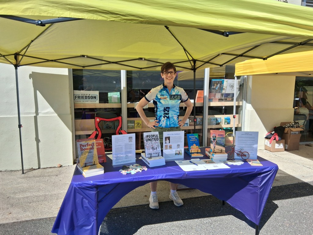 Katherine Pickett standing behind a table displaying books outdoors, under a tent